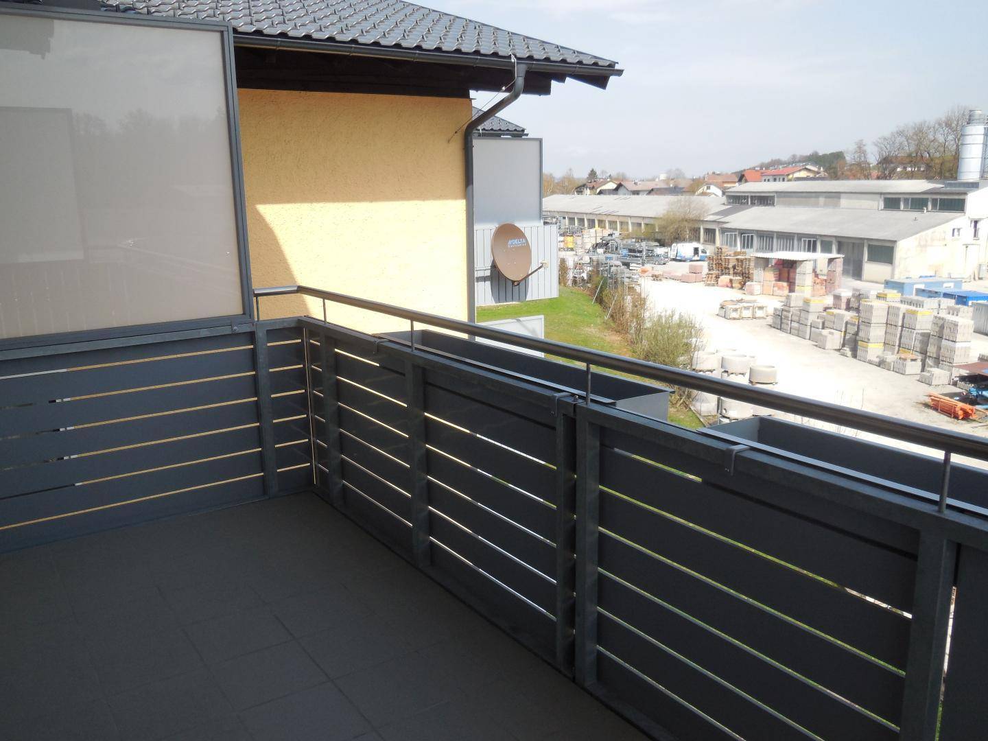 Balkon mit grauen Fliesen und Geländer, bietet einen Blick auf ein Gewerbegebiet mit Gebäuden.