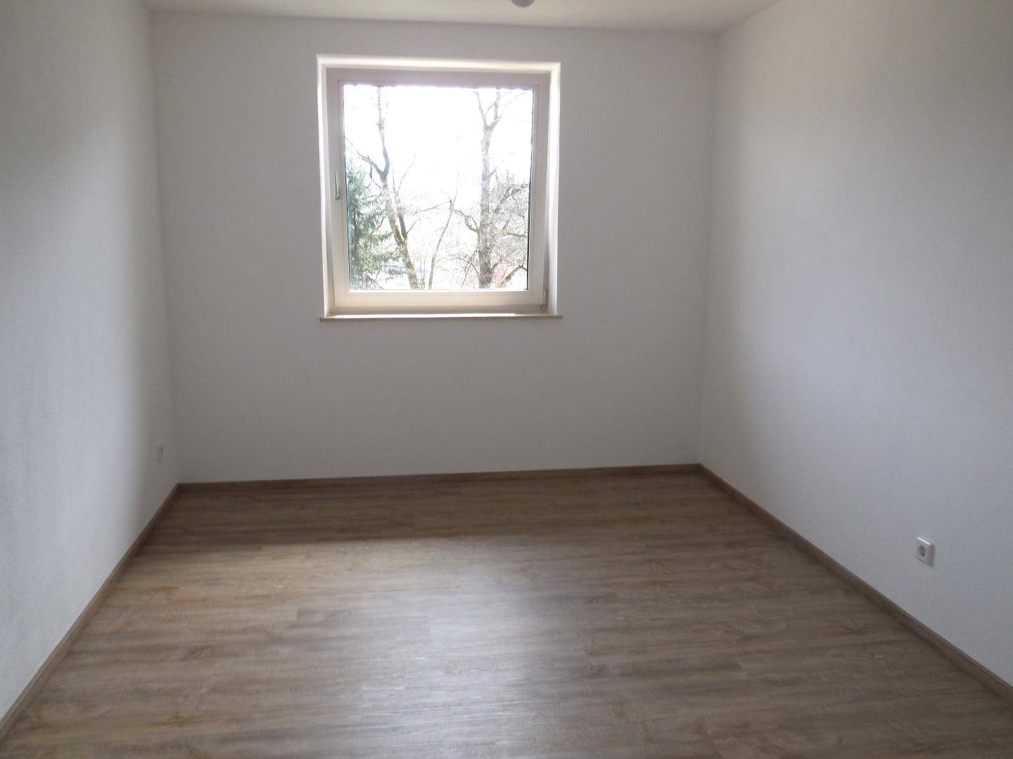 Helles Zimmer mit Laminatboden und einem Fenster, das einen Blick auf die umliegenden Bäume bietet.