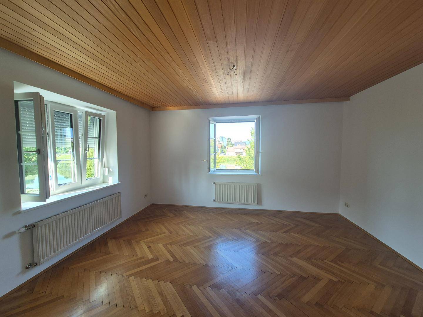 Lichtdurchflutetes Zimmer mit Fischgrätparkett, Holzdecke und weitem Blick ins Grüne.