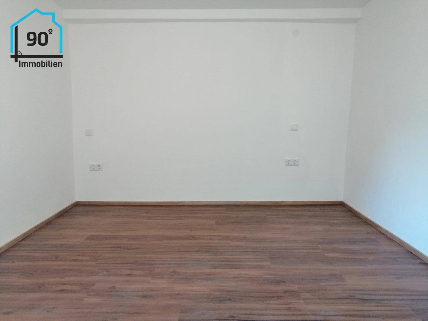 Geräumiger, leerer Raum mit dunklem Laminatboden und weißen Wänden, vielseitig nutzbar und hell.