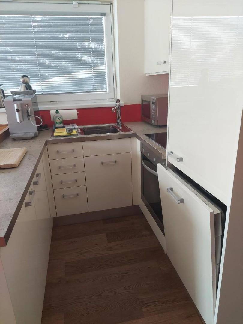 Kleine, funktionale Küche mit weißen Hochglanzschränken, Fenster und integrierten Geräten.