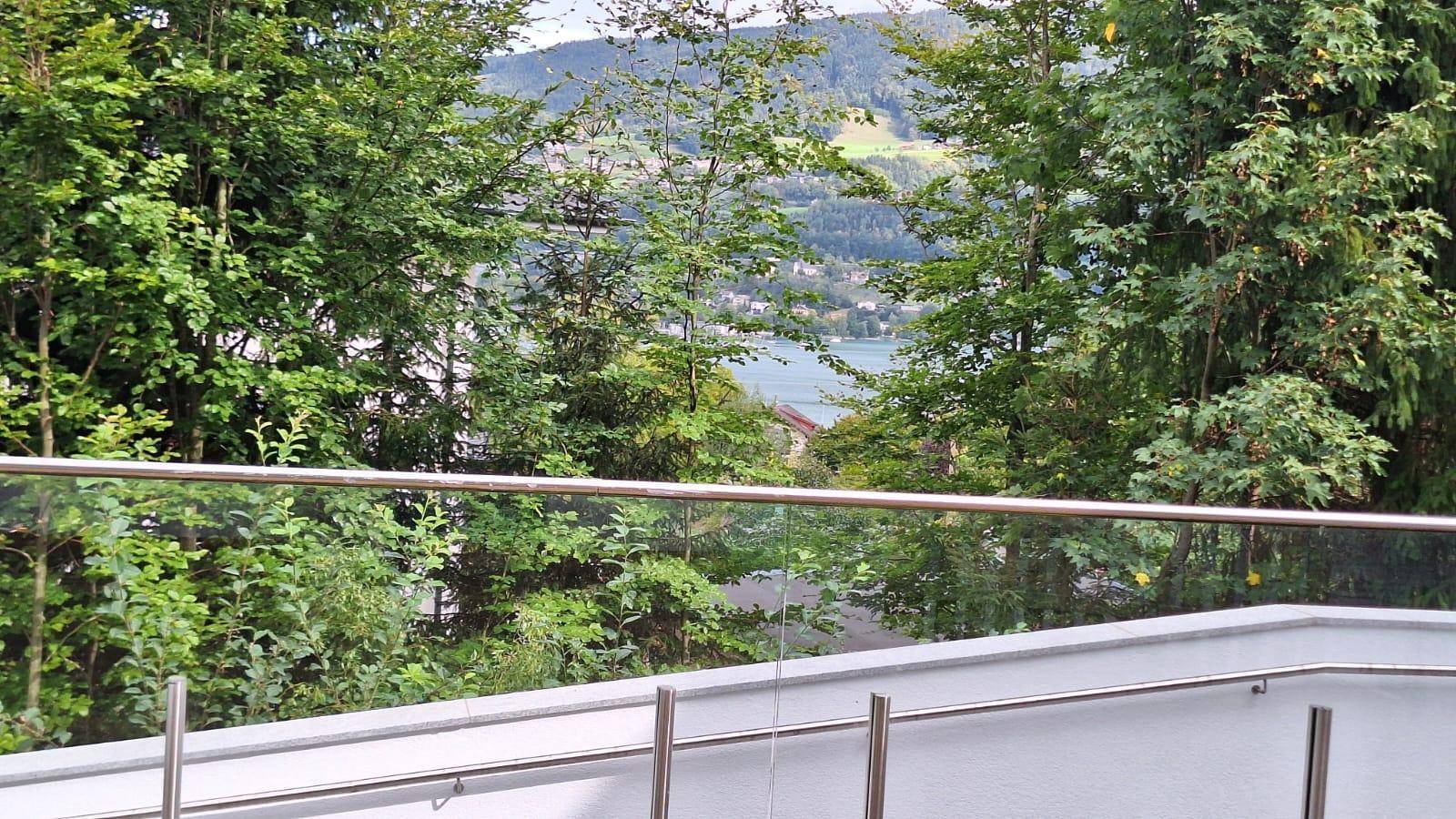 Panoramablick von der Terrasse auf den See und die umliegende Berglandschaft durch grüne Bäume.