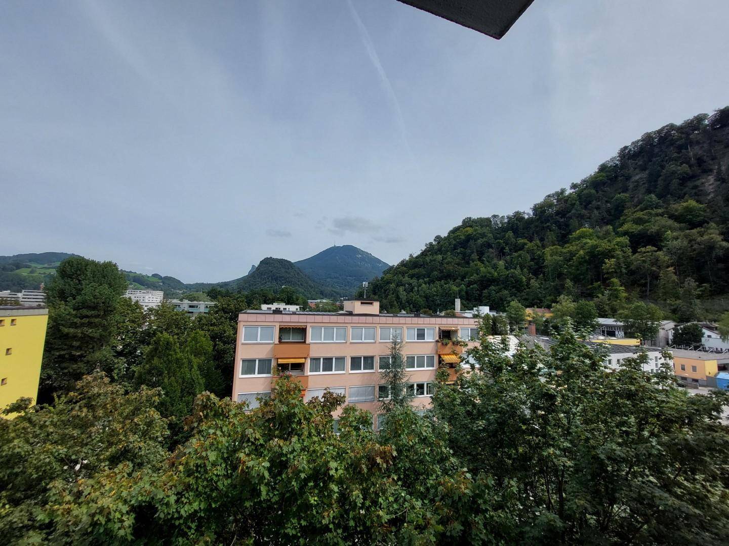 Weitläufiger Blick auf die grüne Umgebung mit Bergen und Wohngebäuden unter bewölktem Himmel.