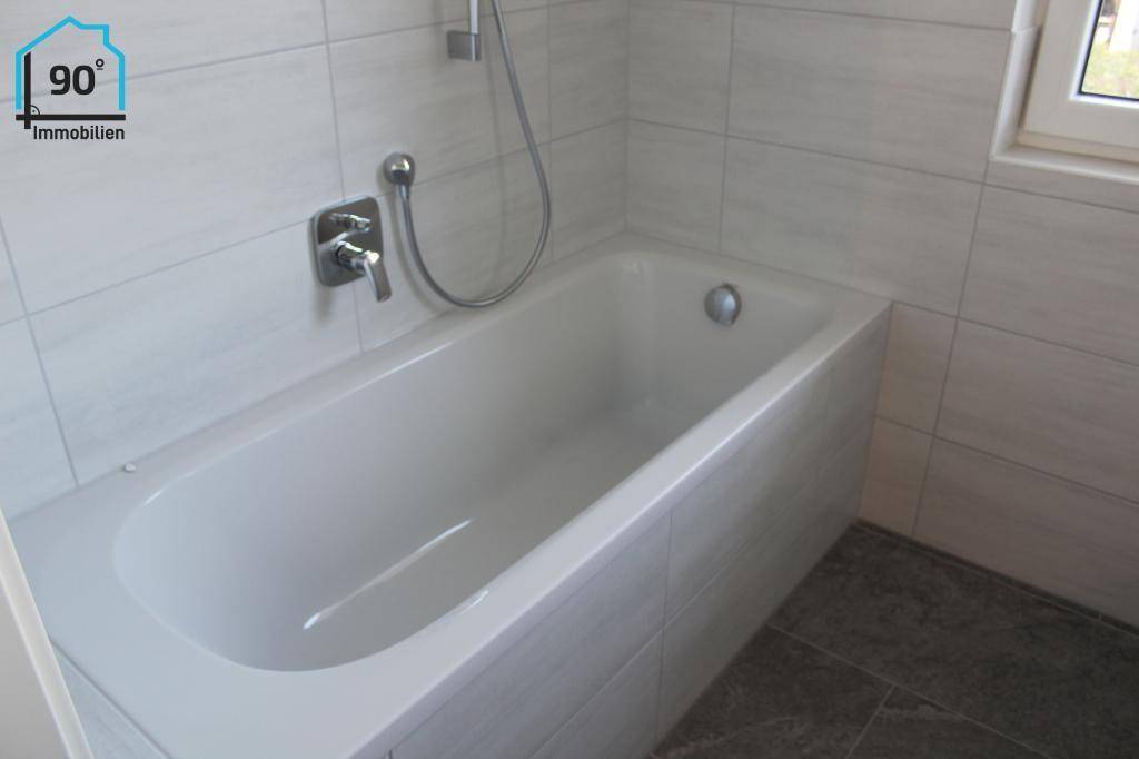 Modernes Badezimmer mit Badewanne, weißen Fliesen und Fenster für Tageslicht.