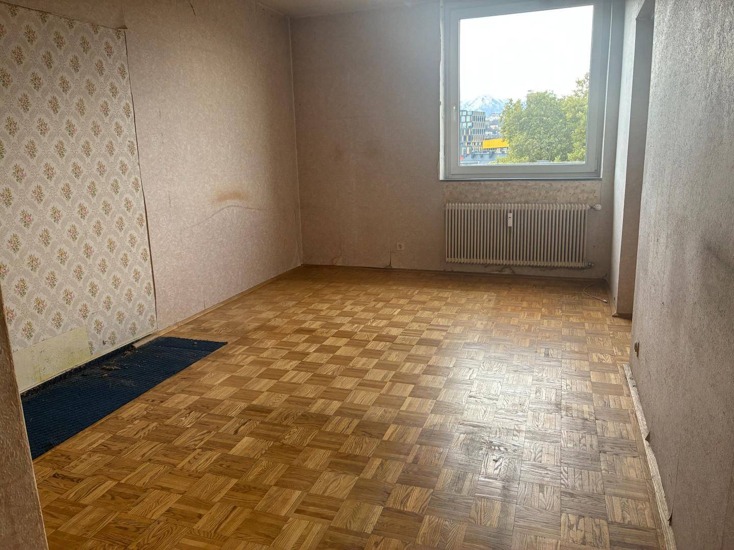 Leeres Zimmer mit Parkettboden und Fenster, das eine Renovierung benötigt.
