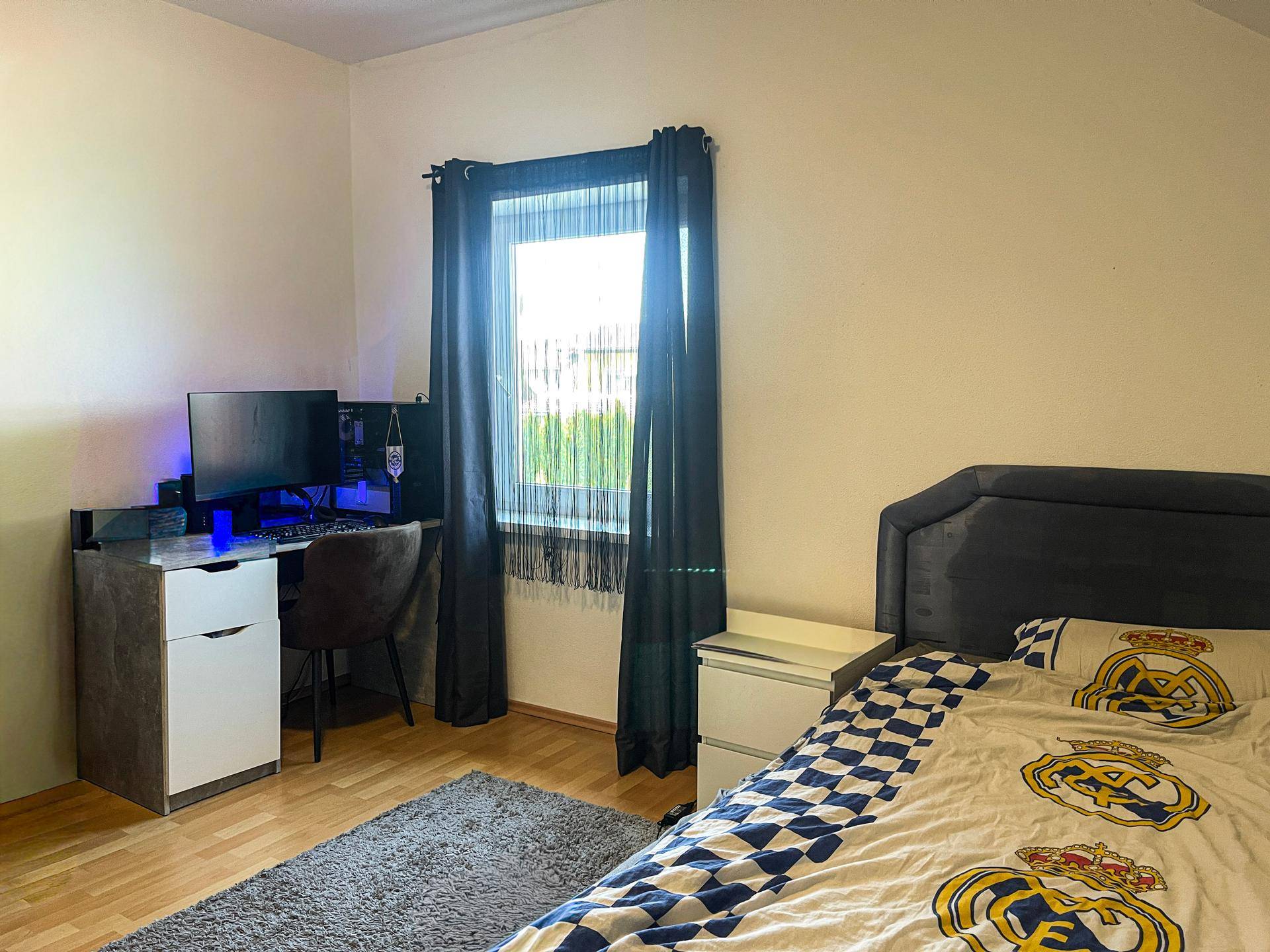 Schlafzimmer mit Bett, Schreibtisch und Fenster, das viel Tageslicht hereinlässt.