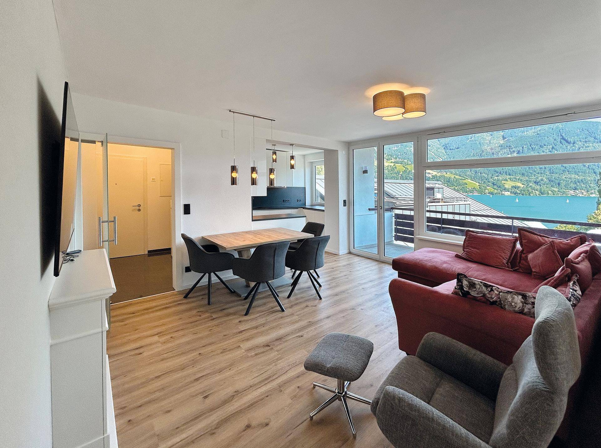 Heller offener Wohn-Essbereich mit Seeblick, rotem Sofa, Esstisch und Holzboden.