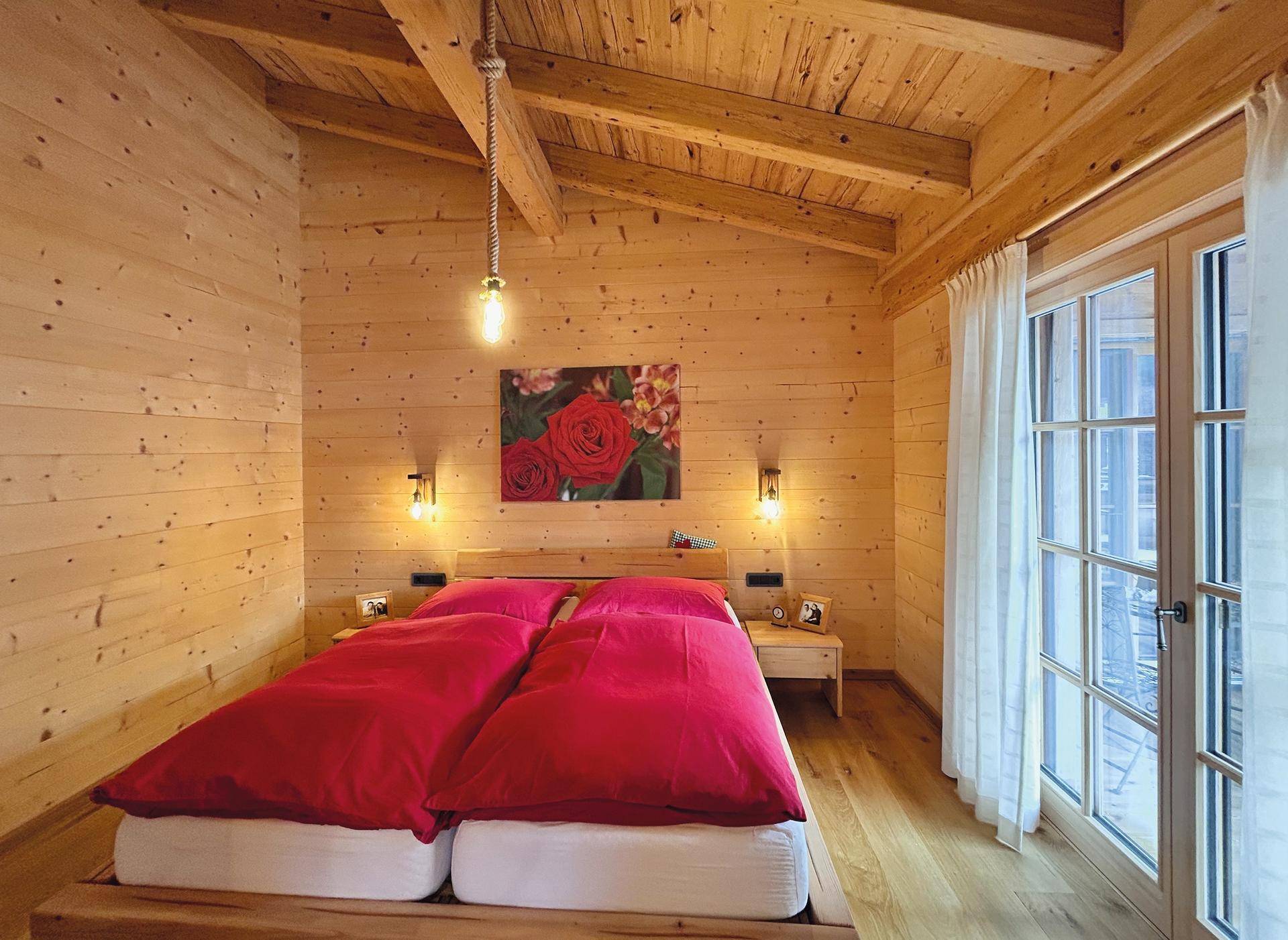 Helles Schlafzimmer mit Holzwänden, Holzbalkendecke und Zugang zum Balkon.