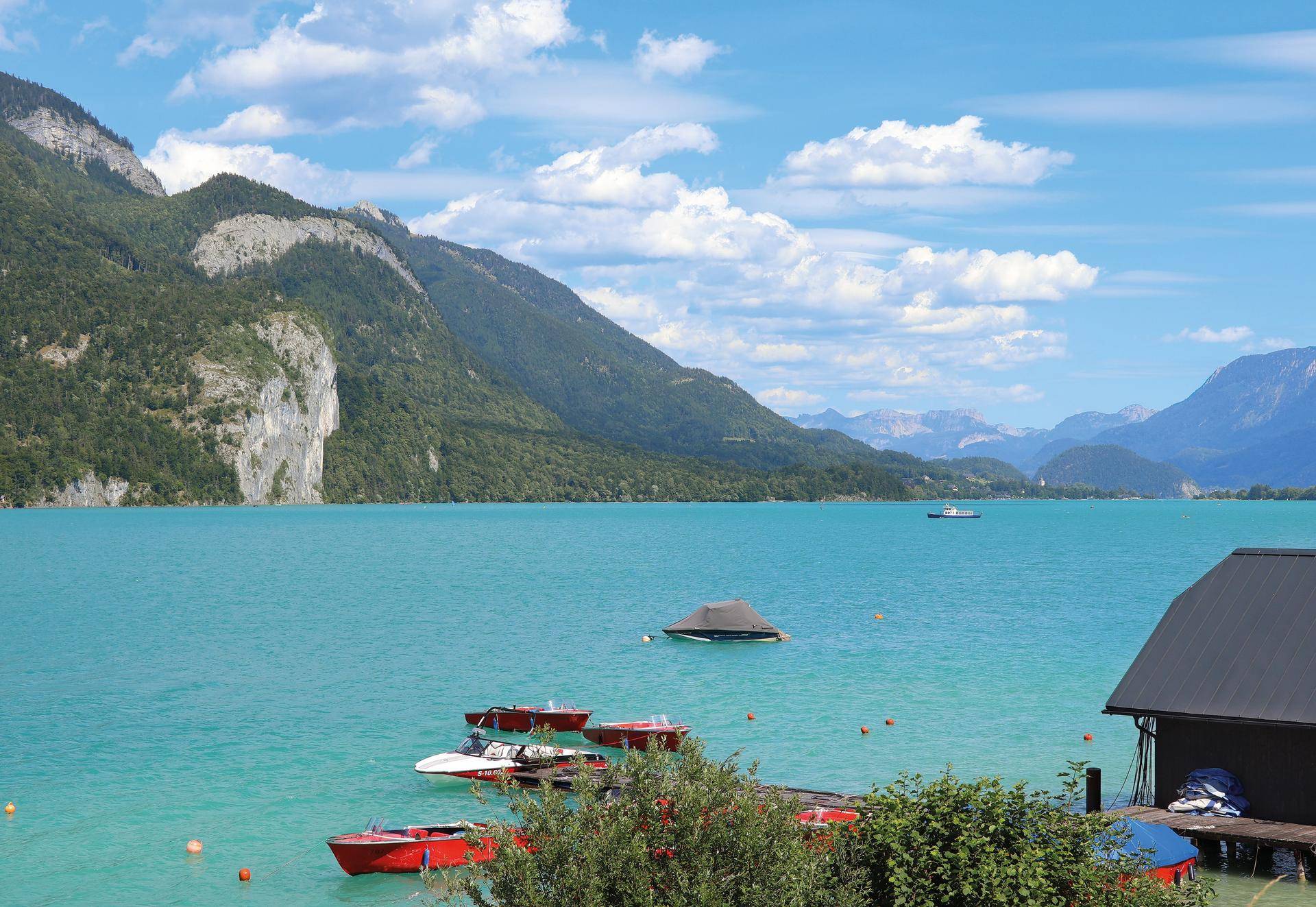 Klarer See mit Booten, umgeben von majestätischen Bergen unter blauem Himmel.