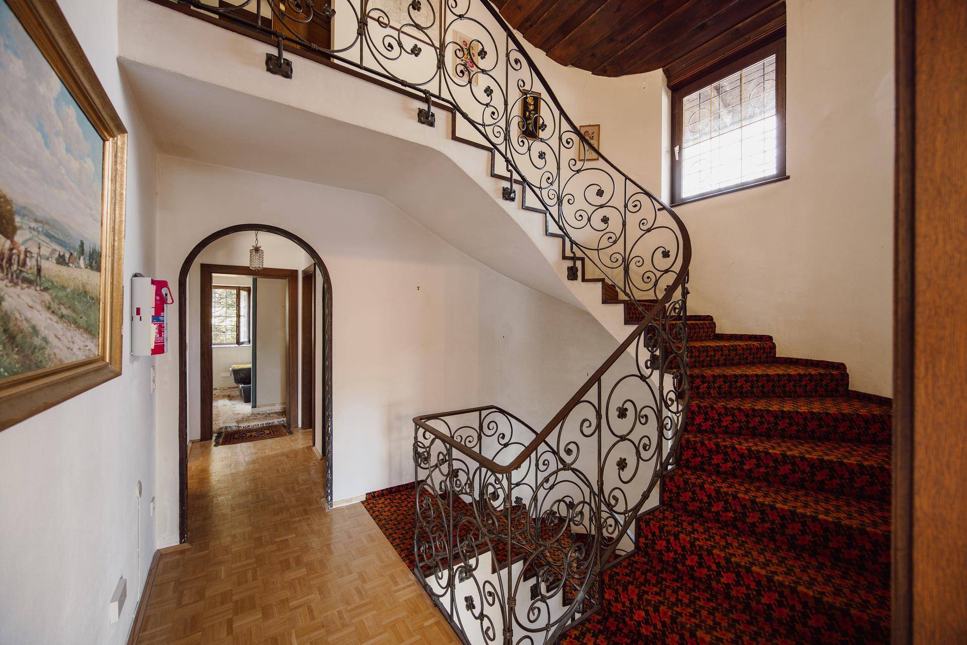 Geschwungene Treppe mit schmiedeeisernem Geländer, gemustertem Teppich und Holzbalkendecke.