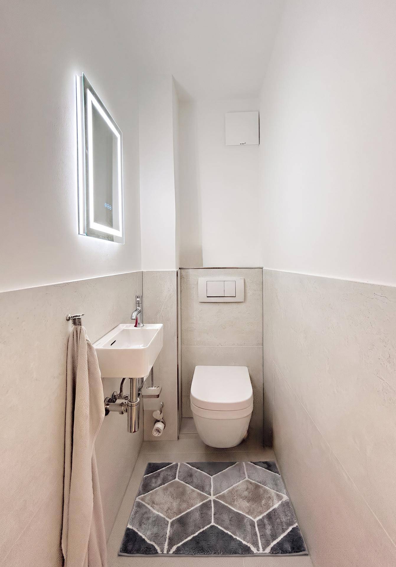 Modernes Gäste-WC mit wandhängender Toilette, kleinem Waschbecken und beleuchtetem Spiegel.