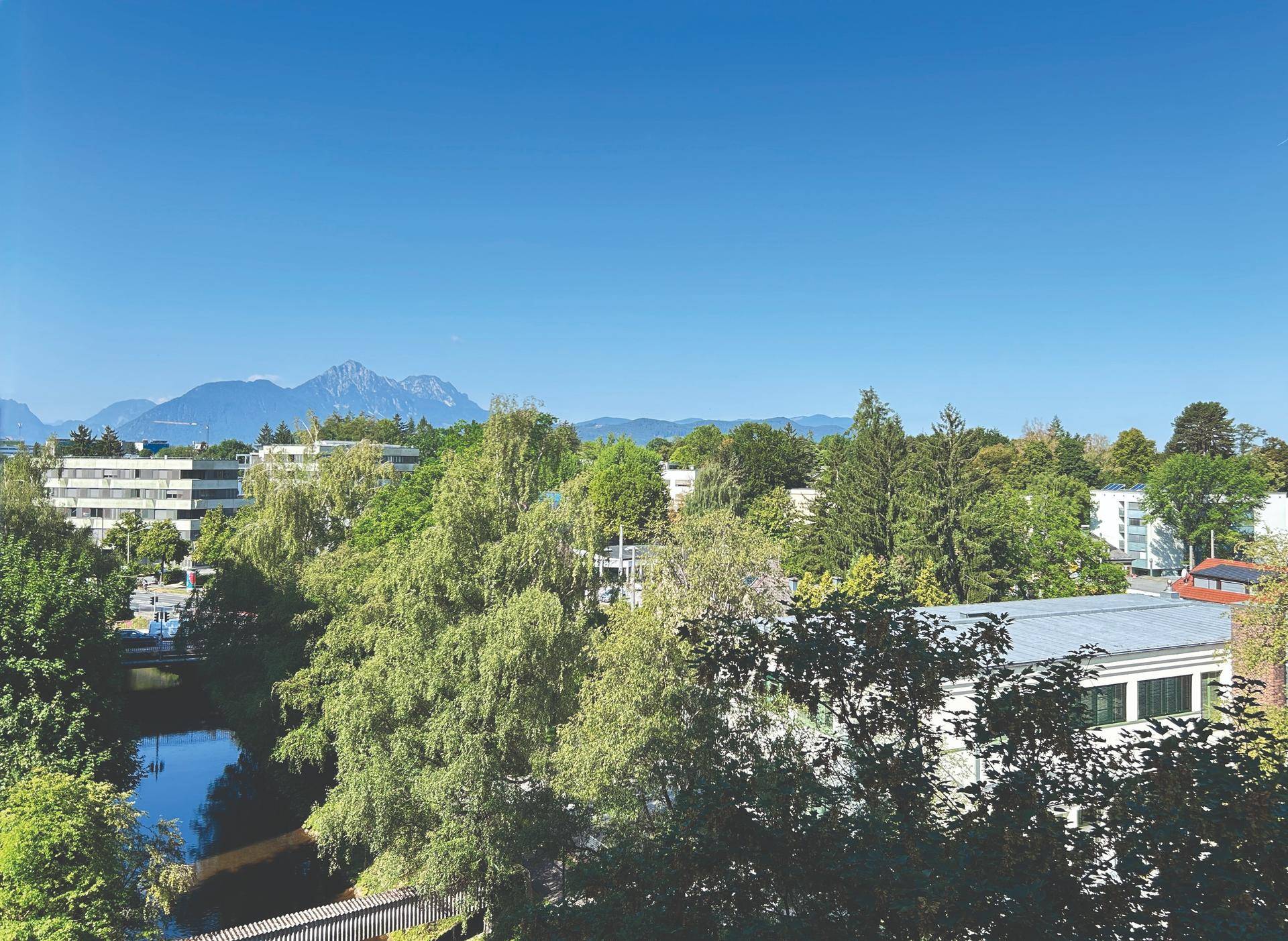 Weitläufiger Blick ins Grüne mit Flusslauf und majestätischen Bergen im Hintergrund.