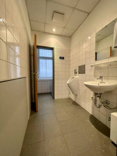 Öffentlicher Waschraum mit weißen Fliesen, Waschbecken, Urinal und Fenster durch eine offene Tür.