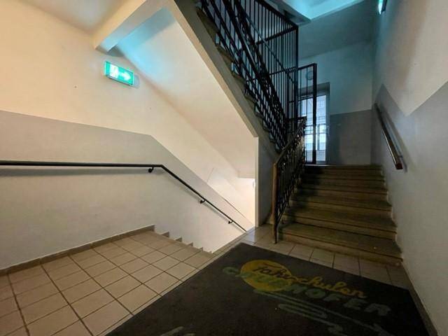 Treppenhaus mit gefliestem Boden, Holzstufen, Metallgeländer und einem Notausgangsschild.