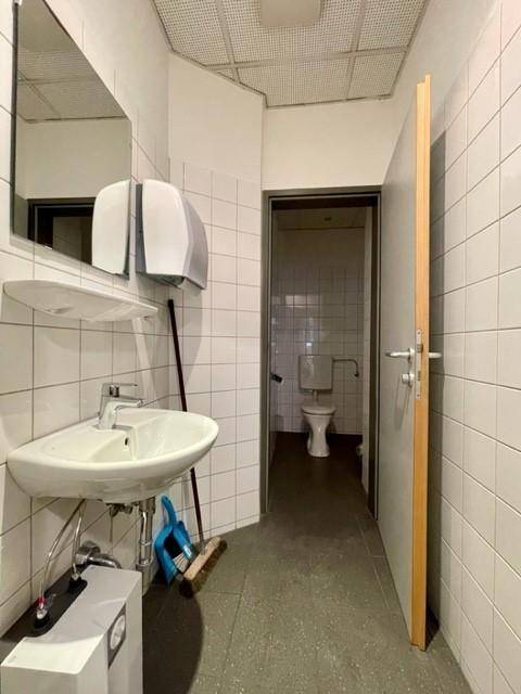 Öffentlicher Waschraum mit weißen Fliesen, Waschbecken und Blick in eine Toilettenkabine.