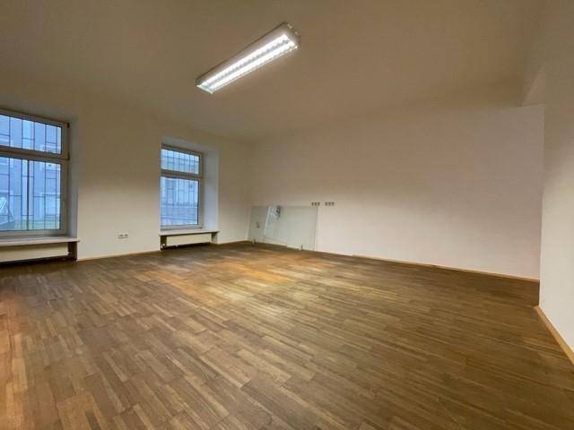 Leerer Büroraum mit Holzfußboden, zwei Fenstern und einer Glasabtrennung im Hintergrund.