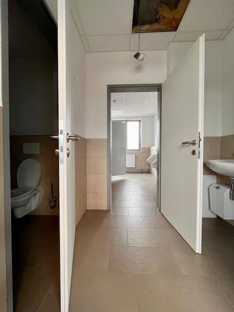 Flur zu den Toilettenräumen mit gefliestem Boden und Blick in einen weiteren Raum.