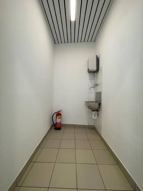 Schmaler Flur oder Abstellraum mit gefliestem Boden, kleinem Waschbecken und einem Feuerlöscher.