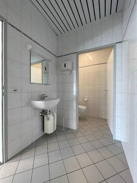 Gefliestes Badezimmer mit Waschbecken, Spiegel und Zugang zu einer separaten Toilettenkabine.