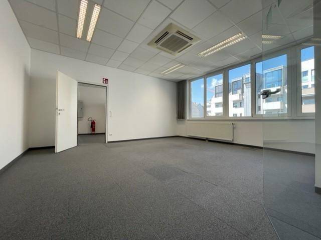 Ein kleineres Büro mit Teppichboden, Heizkörper und einem Fenster, das Tageslicht in den Raum lässt.