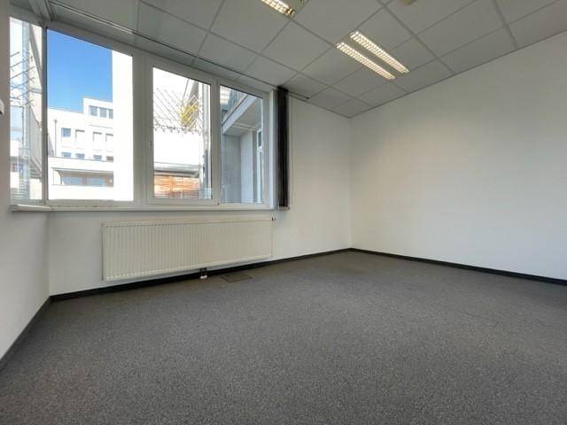 Kompaktes Büro mit Teppichboden, einem großen Fenster und einem Heizkörper, ideal für konzentriertes Arbeiten.
