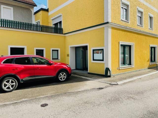 Gelbes Gebäude mit dunklen Fenstern und einem roten Auto davor auf der Straße geparkt.