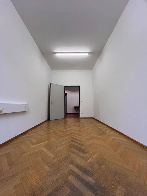Leerer, langer Raum mit Fischgrätparkett, hohen Decken und einer offenen Tür zu einem angrenzenden Bereich.