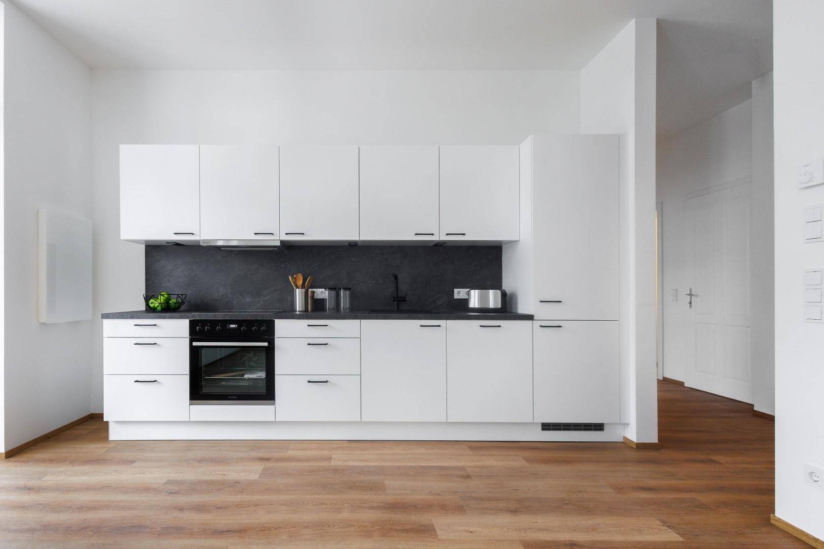 Moderne Einbauküche mit weißen Schränken, dunkler Arbeitsplatte und hellem Holzfußboden.