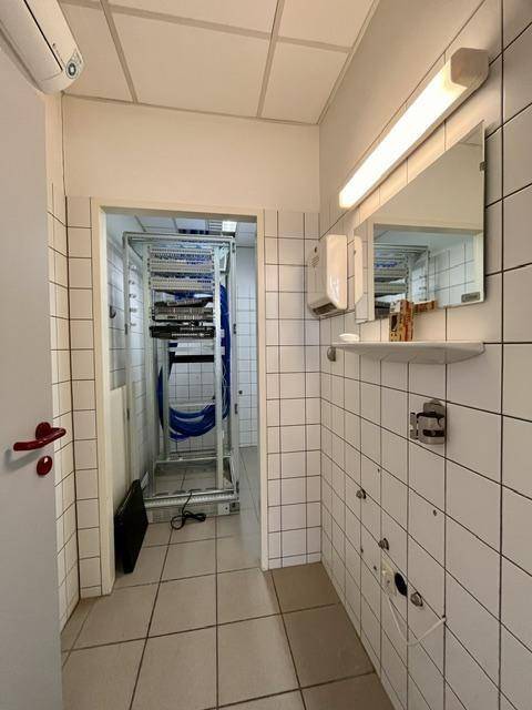 Toilettenbereich mit weißen Fliesen und Zugang zu einem angrenzenden Raum mit Server-Racks oder Lagerregalen.
