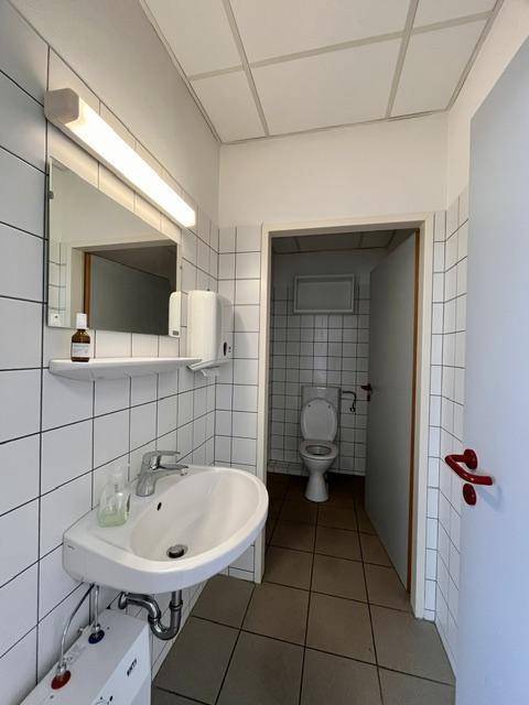 Einfacher Toilettenbereich mit weißen Fliesen, Waschbecken und Spiegel, sowie einer Toilette im Hintergrund.
