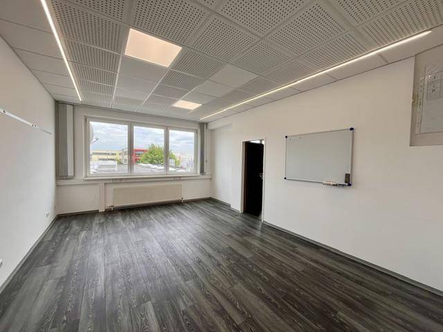 Leerer Büroraum mit großen Fenstern, dunklem Boden und einer Whiteboard-Tafel an der Wand.