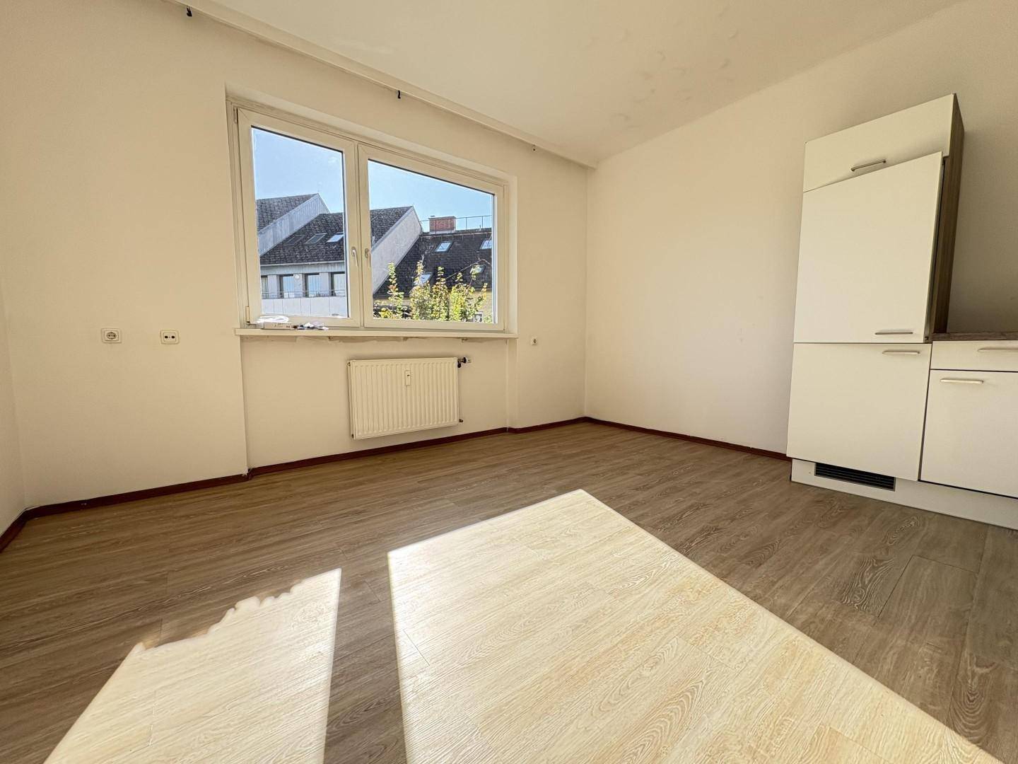 Lichtdurchfluteter Wohnbereich mit großem Fenster und Blick auf die umliegenden Gebäude, ausgestattet mit Laminatboden.