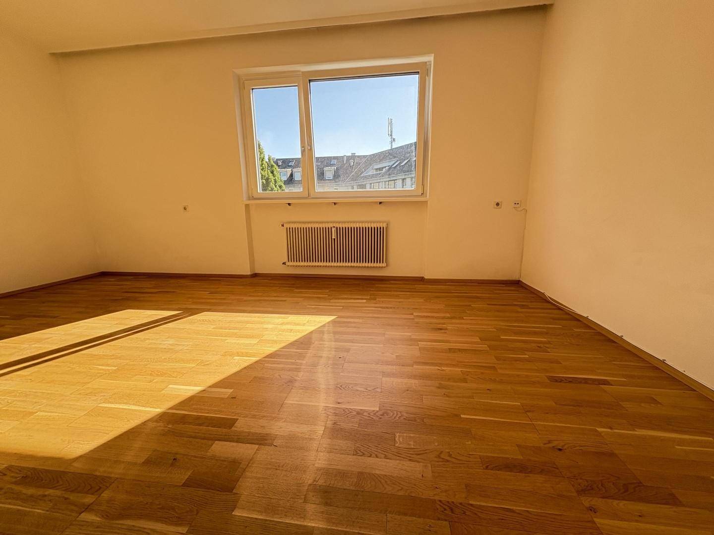 Heller, leerer Raum mit Holzboden und großem Fenster, das viel Tageslicht hereinlässt.