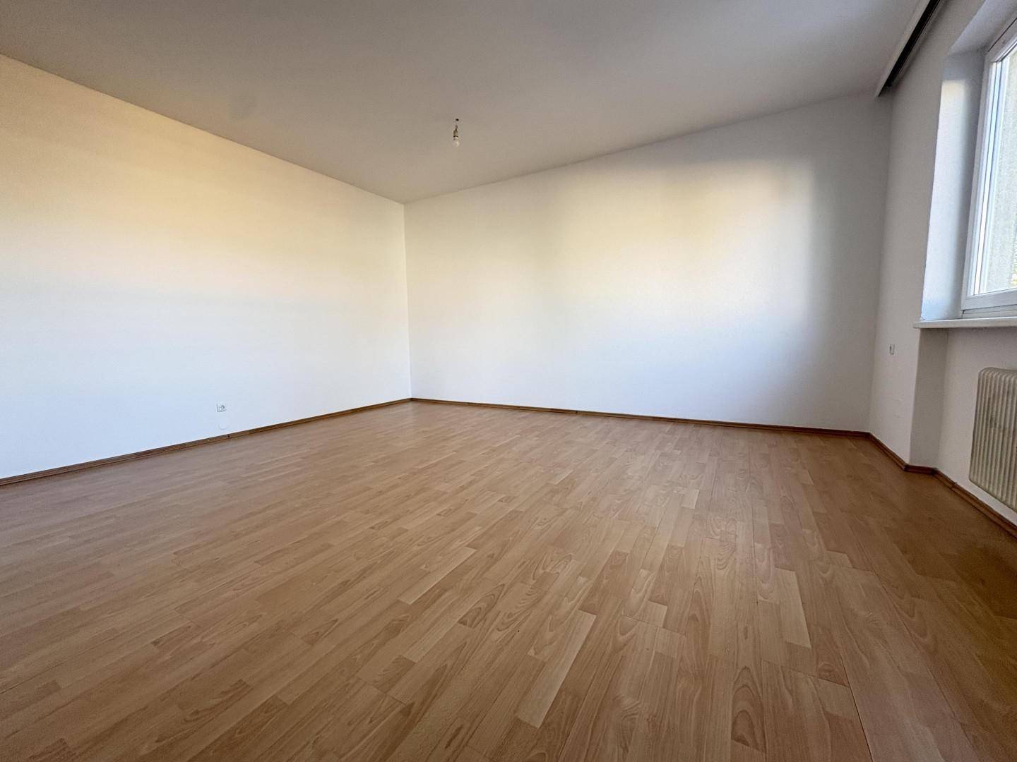 Heller, leerer Raum mit Laminatboden, weißen Wänden und einem Fenster mit Heizkörper.