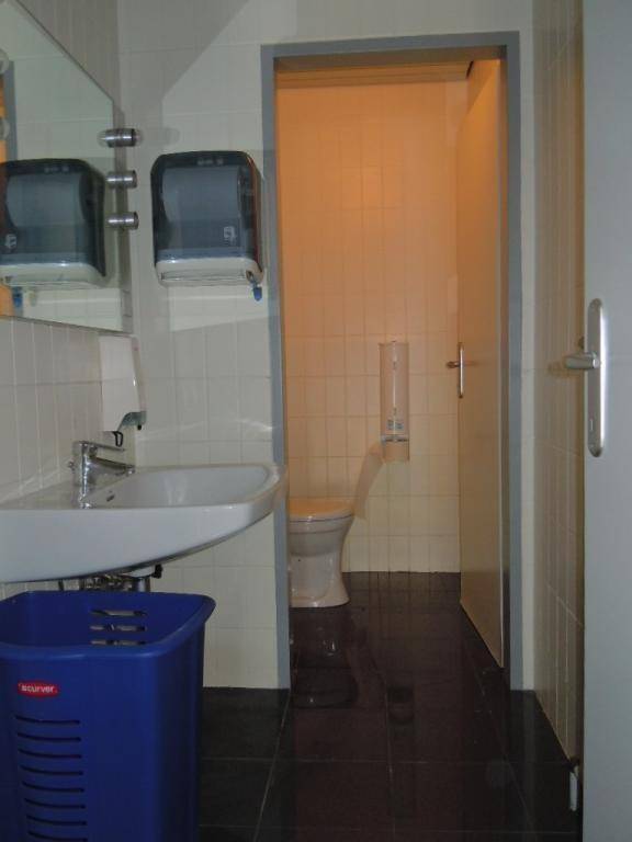Innenansicht einer öffentlichen Toilette mit Waschbecken, Spiegel und einem WC in einem gefliesten Raum.