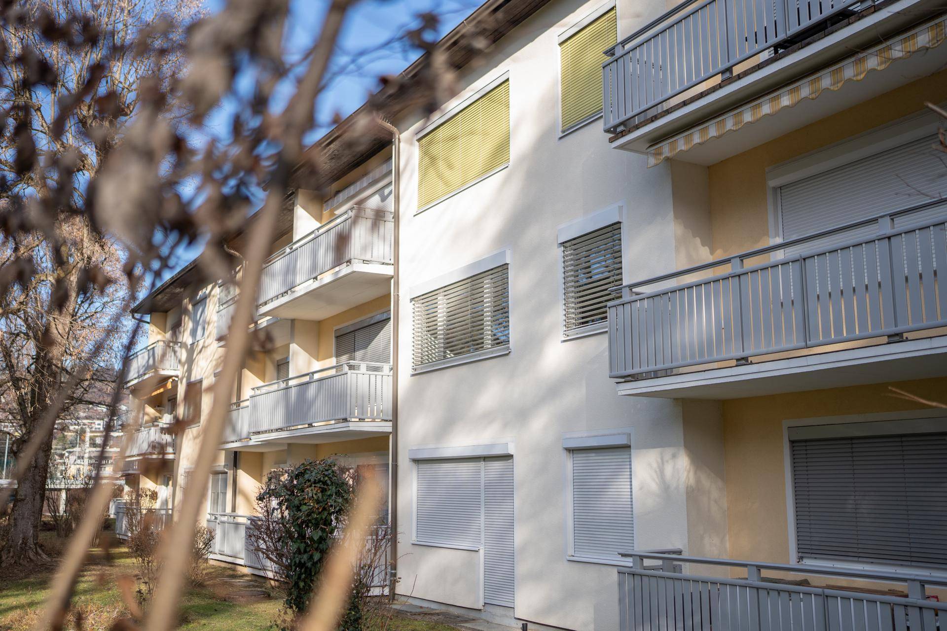 Nahaufnahme der Fassade eines Mehrfamilienhauses mit Balkonen und geschlossenen Jalousien an einem sonnigen Tag.