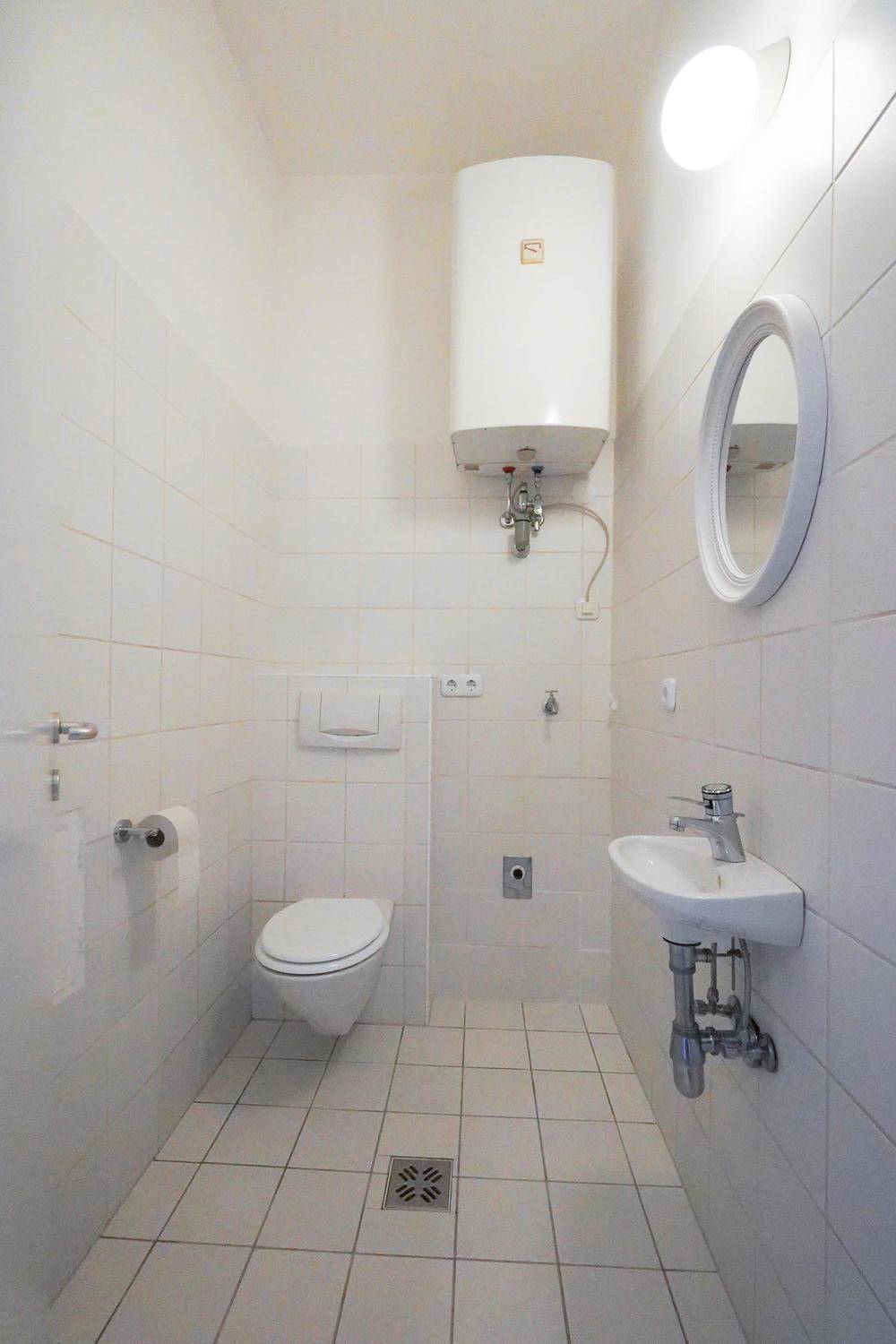 Kleines WC mit wandhängender Toilette, Waschbecken und einem Warmwasserboiler, komplett weiß gefliest.