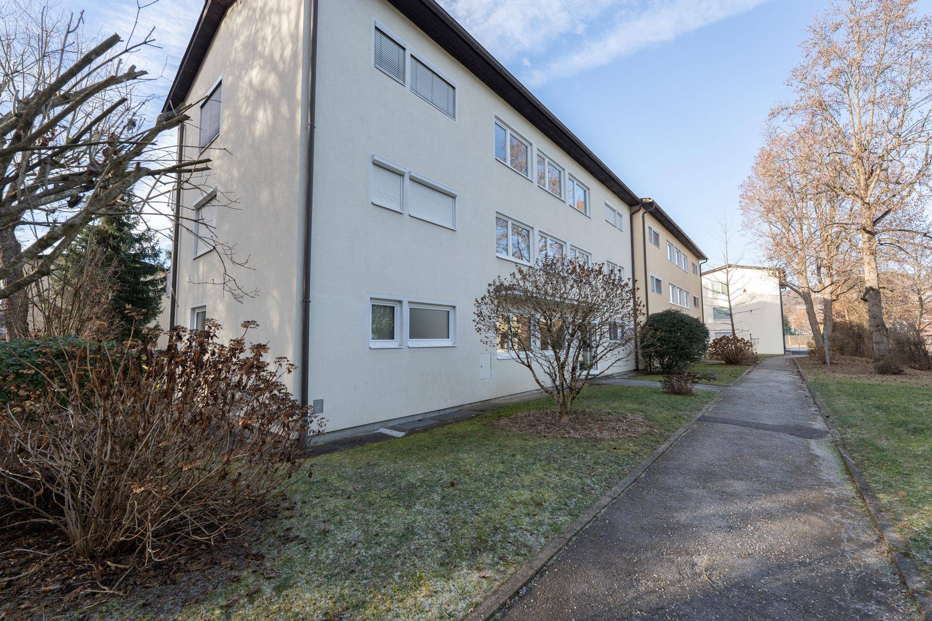 Seitenansicht des Gebäudes mit einem gepflasterten Weg und gepflegten Büschen im Vorgartenbereich.