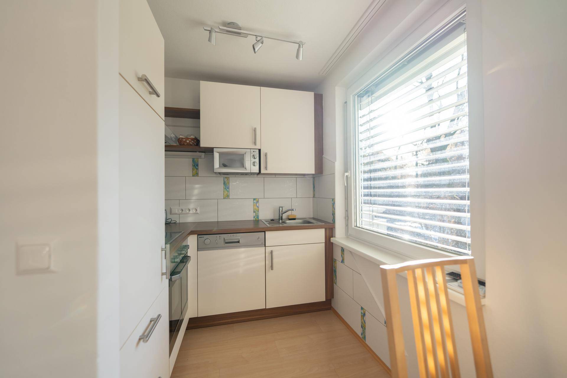 Kompakte Küche mit weißen Schränken, Holztheke und Fenster mit Jalousien.