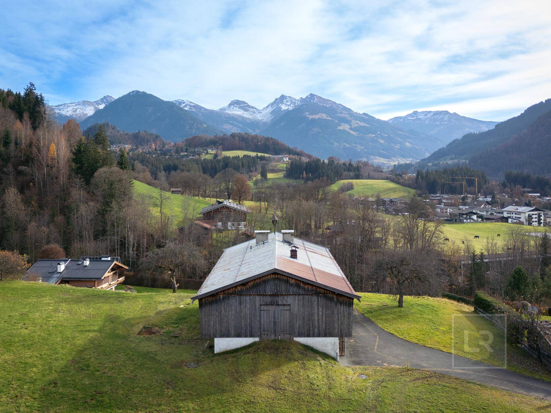 Rustikales Holzgebäude mit Satteldach, umgeben von grünen Wiesen und einer beeindruckenden Bergkette.