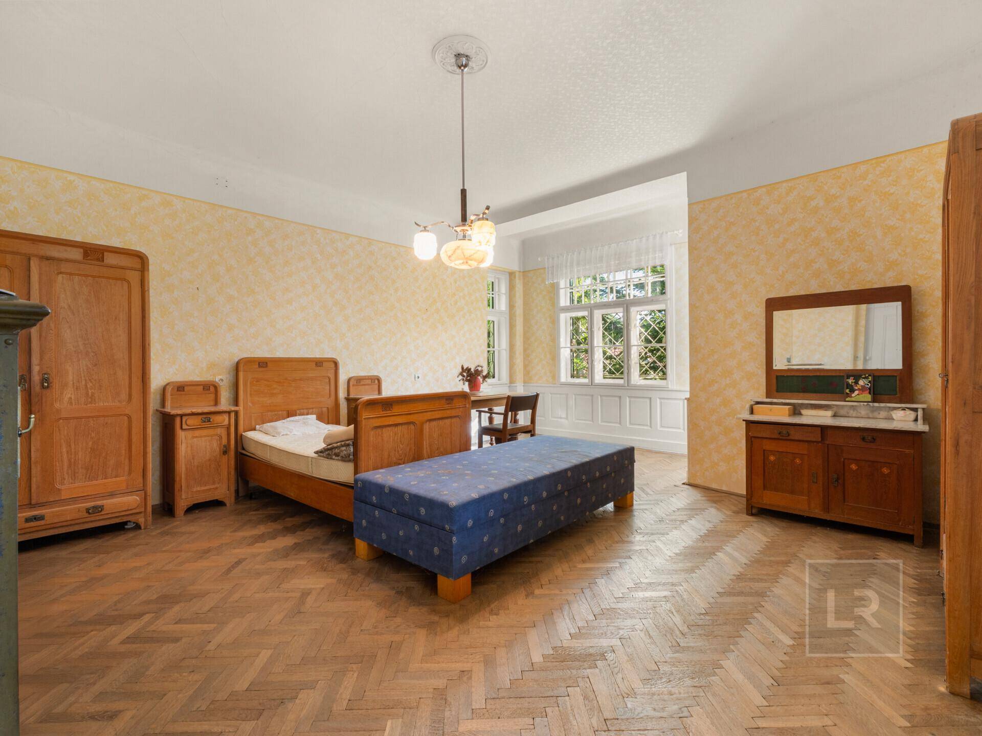 Geräumiges Schlafzimmer mit Fischgrätparkett, antiken Holzmöbeln und gelber Mustertapete.