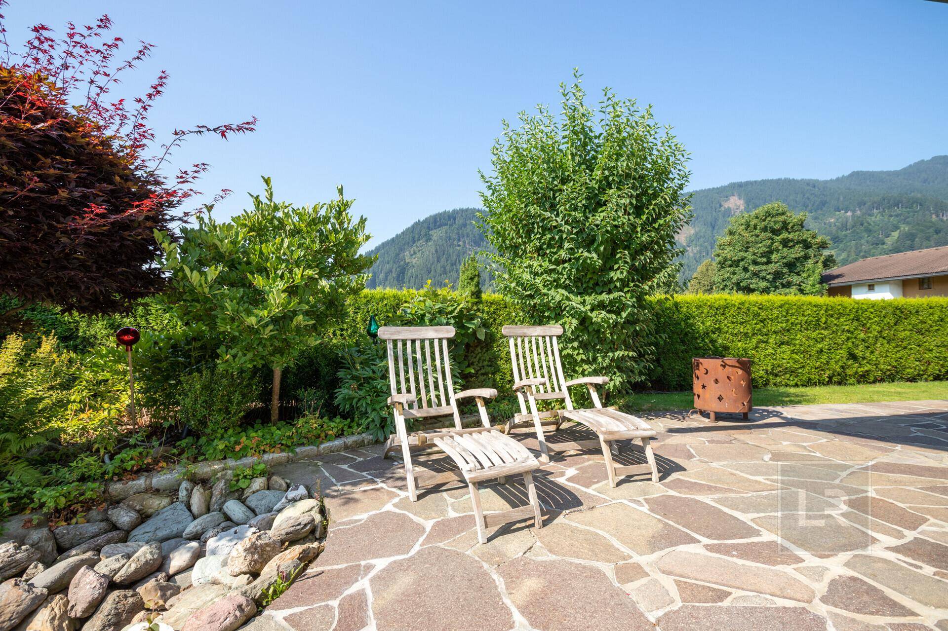 Sonniger Terrassenbereich mit Natursteinplatten und Holzstühlen, umgeben von Hecken und Bäumen.