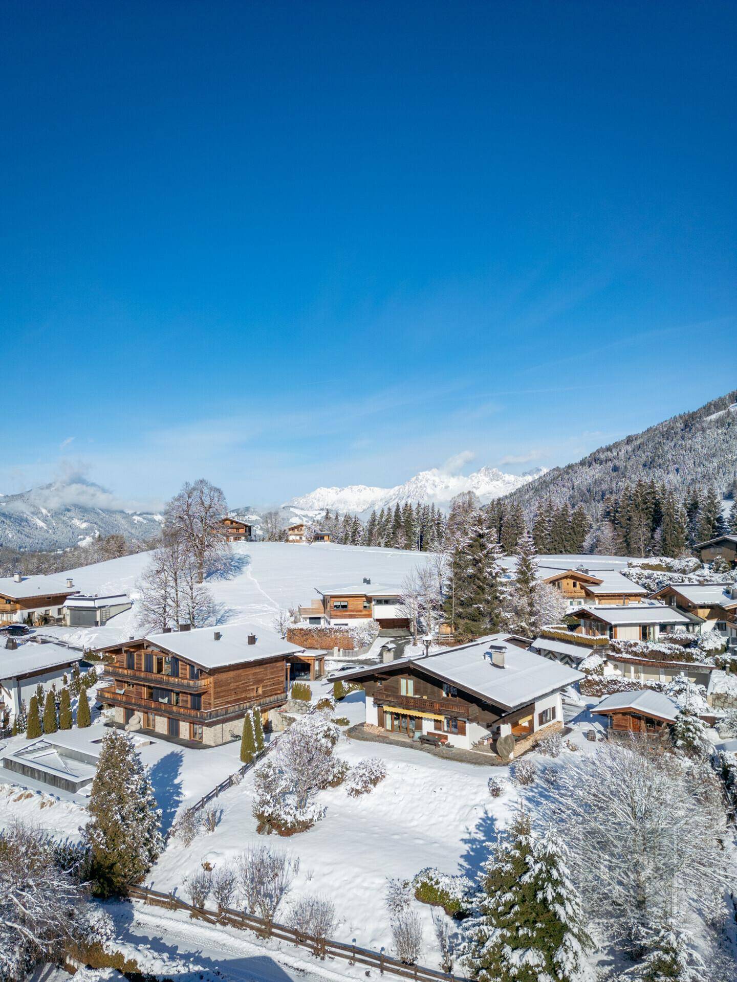 Weitläufige Ansicht einer Chalet-Siedlung in einer verschneiten Bergregion mit klarem, blauem Himmel.
