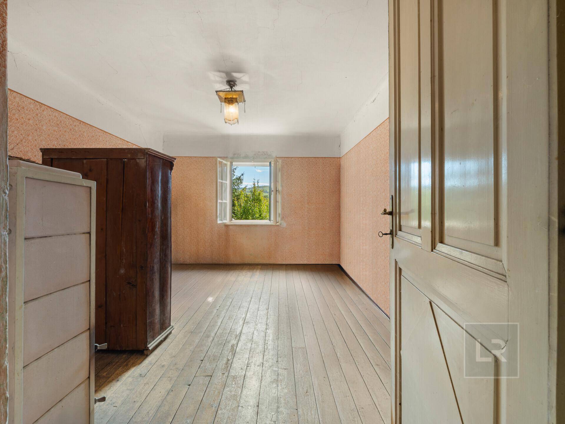 Leeres Zimmer mit Dielenboden, gemusterter Tapete und einem Fenster mit Ausblick ins Grüne.