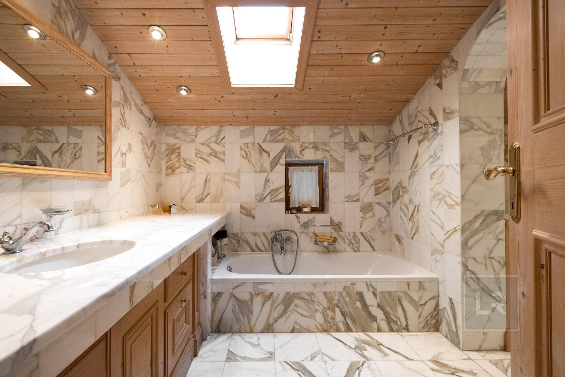 Marmorbad mit Badewanne, Doppelwaschbecken und Holzdecke, durch ein Dachfenster hell beleuchtet.