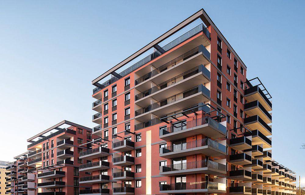 Modernes Wohngebäude mit zahlreichen Balkonen und einer markanten orangefarbenen Fassade unter klarem Himmel.