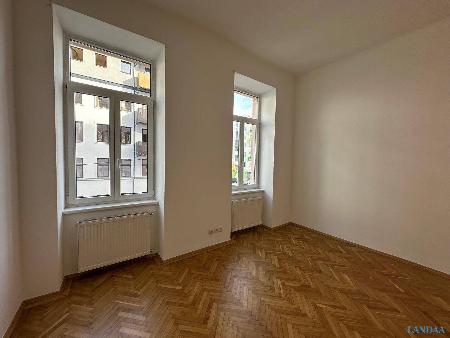 Geräumiger, heller Raum mit klassischem Fischgrätparkett und großen Fenstern zur Straße.