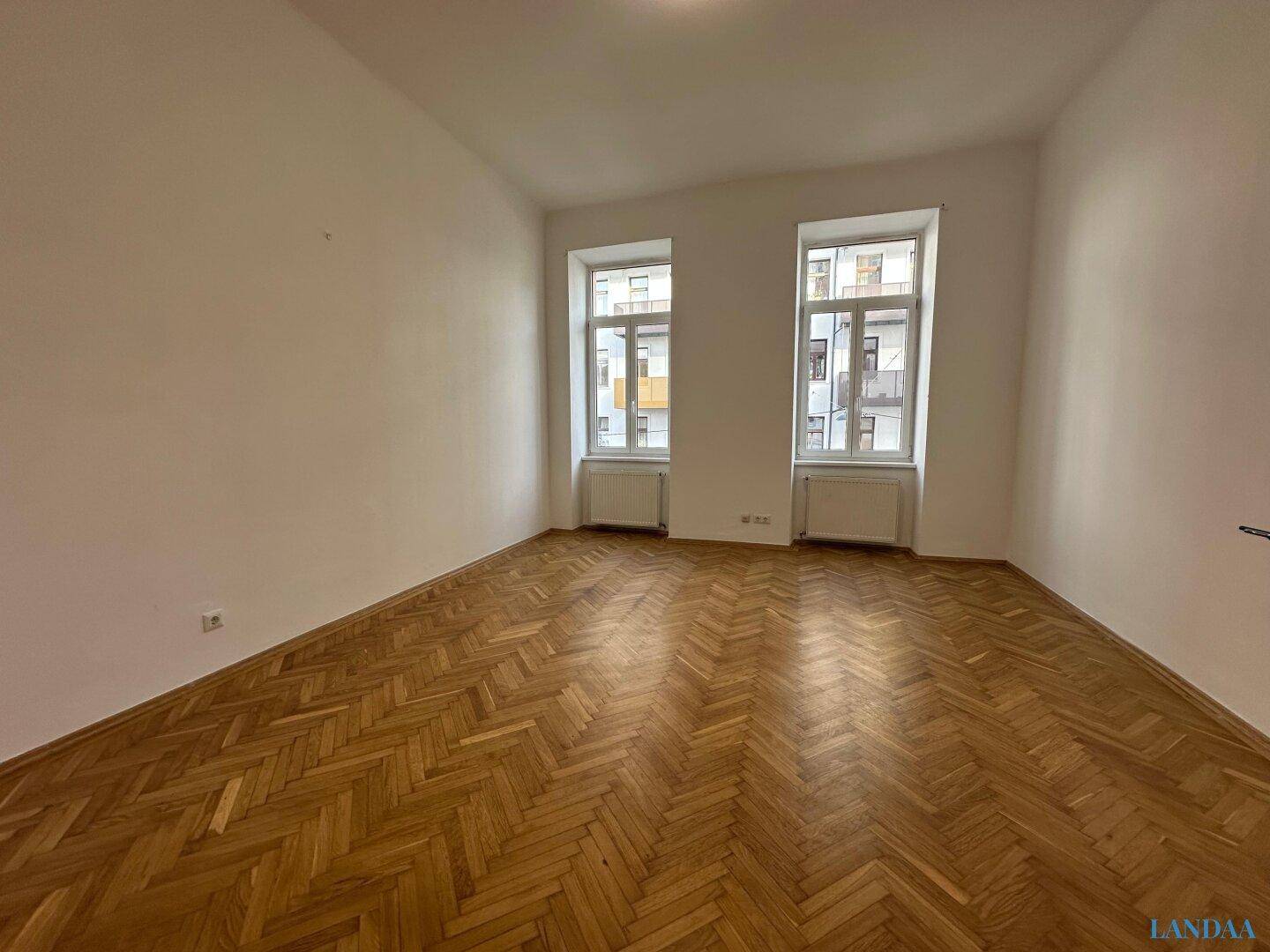 Heller Raum mit Fischgrätparkett und zwei Fenstern, ideal als Wohn- oder Schlafzimmer.