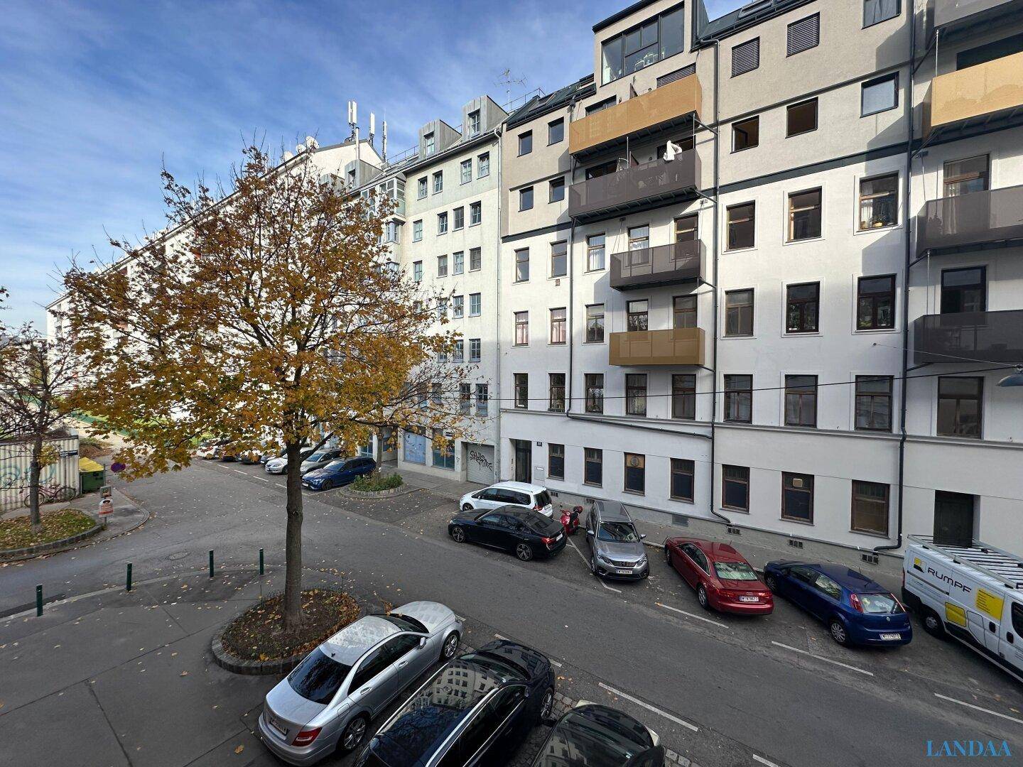 Blick auf die Straße mit parkenden Autos und umliegenden Wohngebäuden unter blauem Himmel.