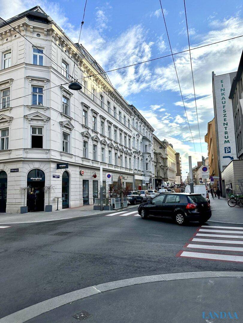 Belebte Straßenszene mit einem Eckgebäude im klassischen Stil und vorbeifahrenden Autos.